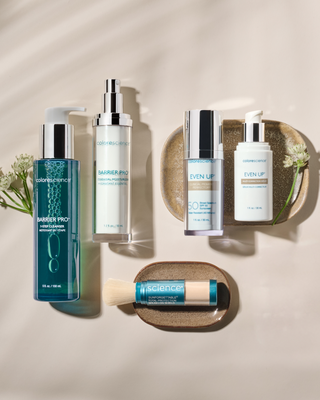 Flat lay of five Colorescience skincare products arranged neatly on a light beige surface with two small tan ceramic trays and delicate white flowers. Products include BARRIER PRO™ Total Treatment Cleanser, BARRIER PRO™ Essential Moisturizer, EVEN UP® Clinical Pigment Perfector SPF 50, EVEN UP® Multi-Correction Serum, and a brush-on Sunforgettable® Total Protection™ product in a turquoise container.