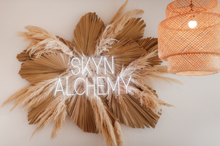 Skyn Alchemy logo surrounded by a decorative arrangement of dried palm leaves and pampas grass, with a woven light fixture on the right, creating a warm and natural aesthetic.
