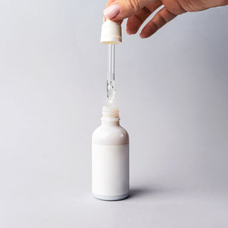 Hand holding a dropper above a minimal white serum bottle, showcasing a skincare product designed for hydration, nourishment, and a glowing complexion.