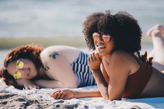 Two women lounging on the beach in sunglasses and swimsuits, highlighting the importance of zinc-based sunscreen for sun protection and healthy skin—your skincare staple during National Skin Cancer Awareness Month.