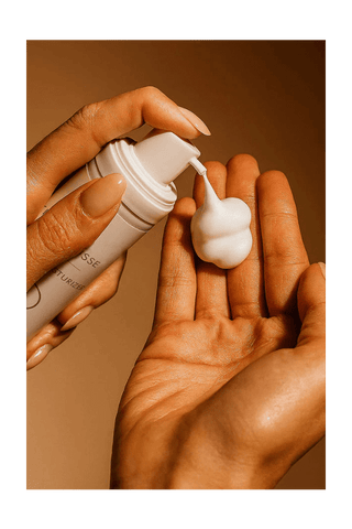 Hand applying cream from a tube onto another hand against a brown background