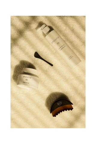 Epicutis body care set arranged on a textured cream surface with soft shadow overlays, including a pump bottle labeled body treatment, a small jar labeled body scrub, a black spatula, and a dark brown wooden massage brush.