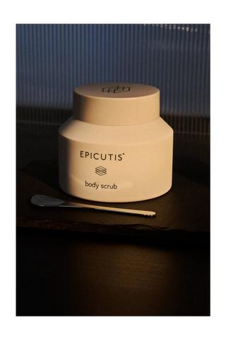 A cream-colored jar of EPIcutis Body Scrub with a matching lid, placed on a dark wooden surface next to a metal spatula, set against a dark blue ribbed background.