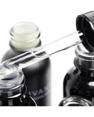 Close-up of Vivant Serum clear glass dropper holding a transparent liquid above black cosmetic bottles, with one bottle showing partial white branding text on a white background.