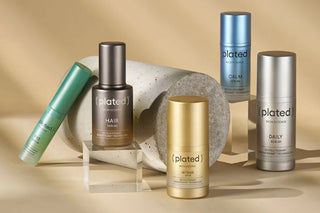 Aesthetic display of Plated Skin Science products, including serums in metallic green, bronze, gold, silver, and blue packaging, arranged around a stone cylinder on a beige background.